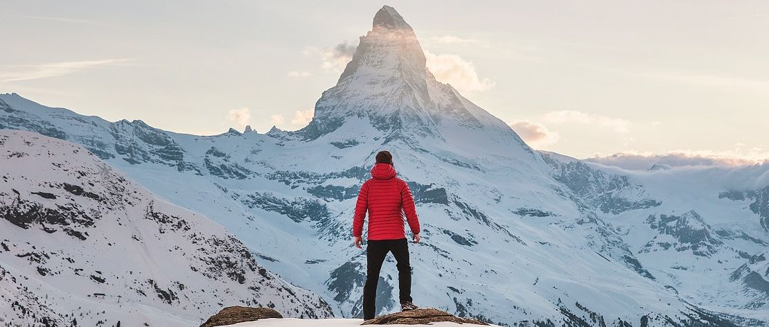 Ein Mann steht vor einem hohen schneebedeckten Berg. Er blickt in Richtung der Bergspitze.