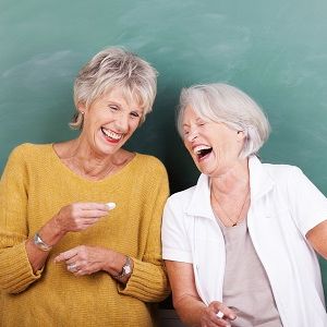 Zwei ältere Lehrerinnen mit grauen Haaren stehen vor einer Schultafel und lachen herzlich.