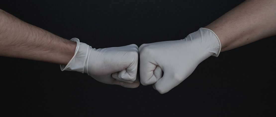 Zwei Personen mit medizinischen Handschuhen begrüßen sich mit einer Geste, bei der sie ihre Fäuste kurz gegeneinanderdrücken.